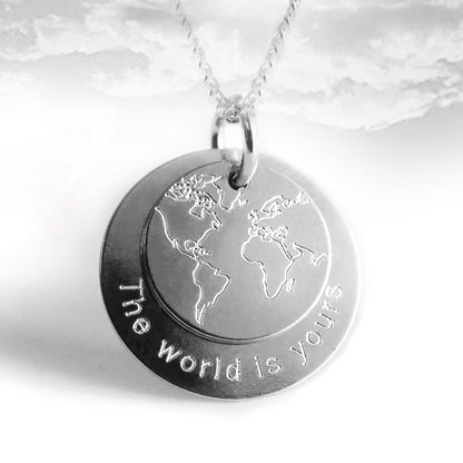 Weltkarten-Kette aus Silber mit zwei Anhängern unterschiedlicher Größe. Der kleinere Anhänger zeigt eine Weltkarte, der größere Anhänger trägt am Rand die Gravur „the world is yours“.